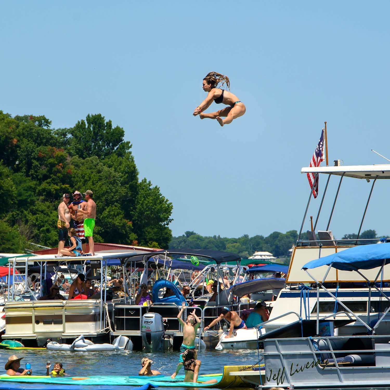 Woman mid-air in a tuck jump cliff-jumping over pontoon boats in a Grand Lake cove